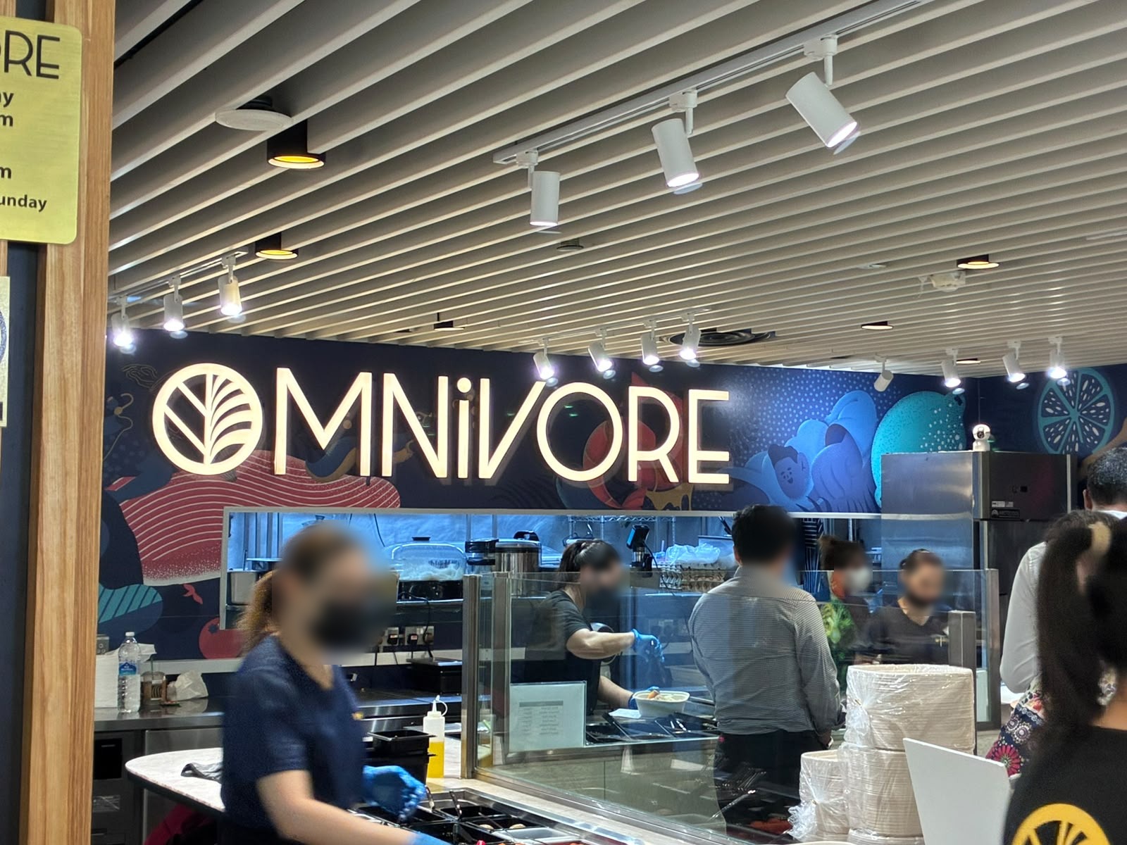 Omnivore counter and mural signage at The Basement food hall, Singapore (faces blurred)