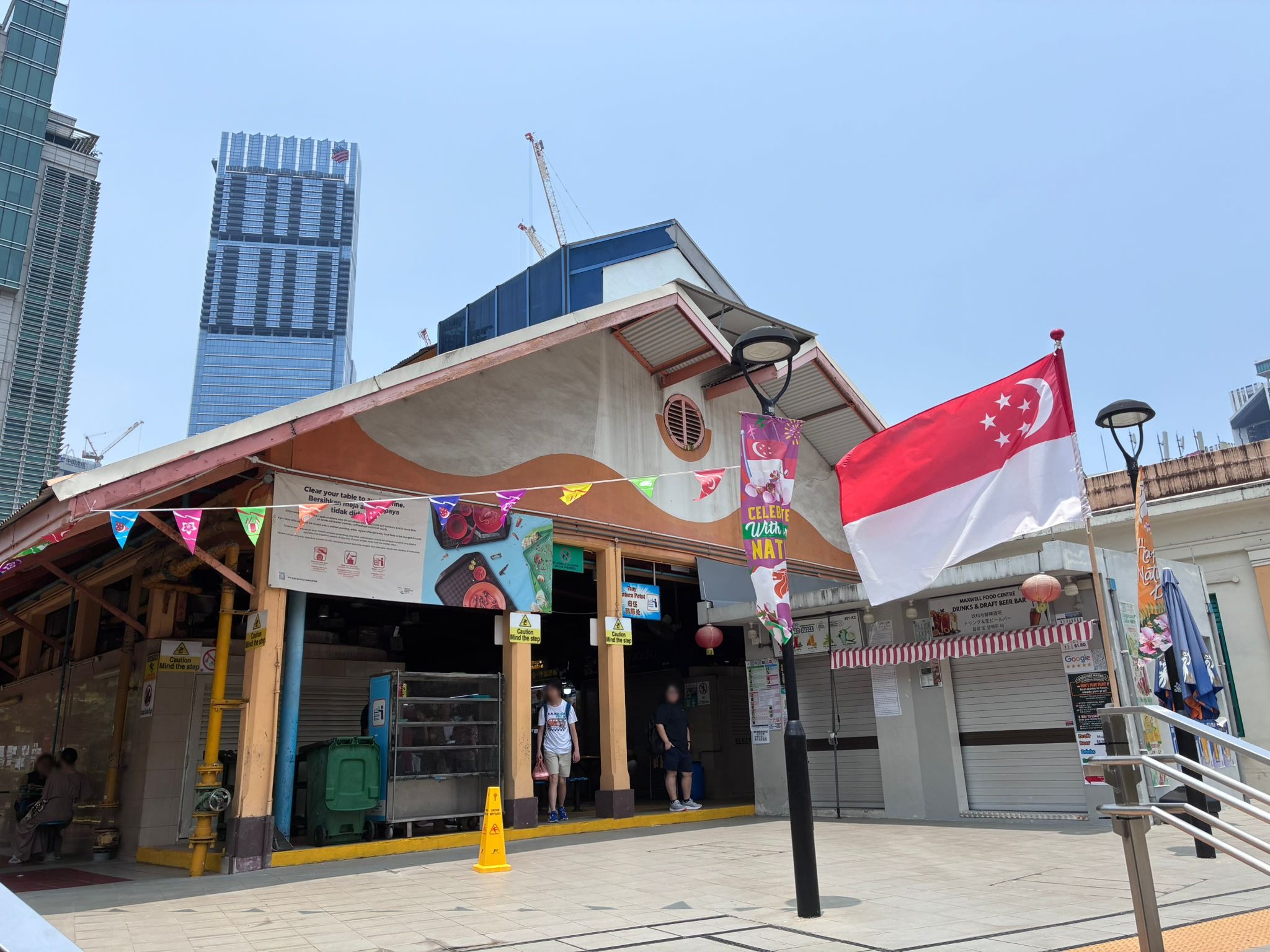 Maxwell Food Centre exterior with Singapore flag and national day bunting (faces blurred)
