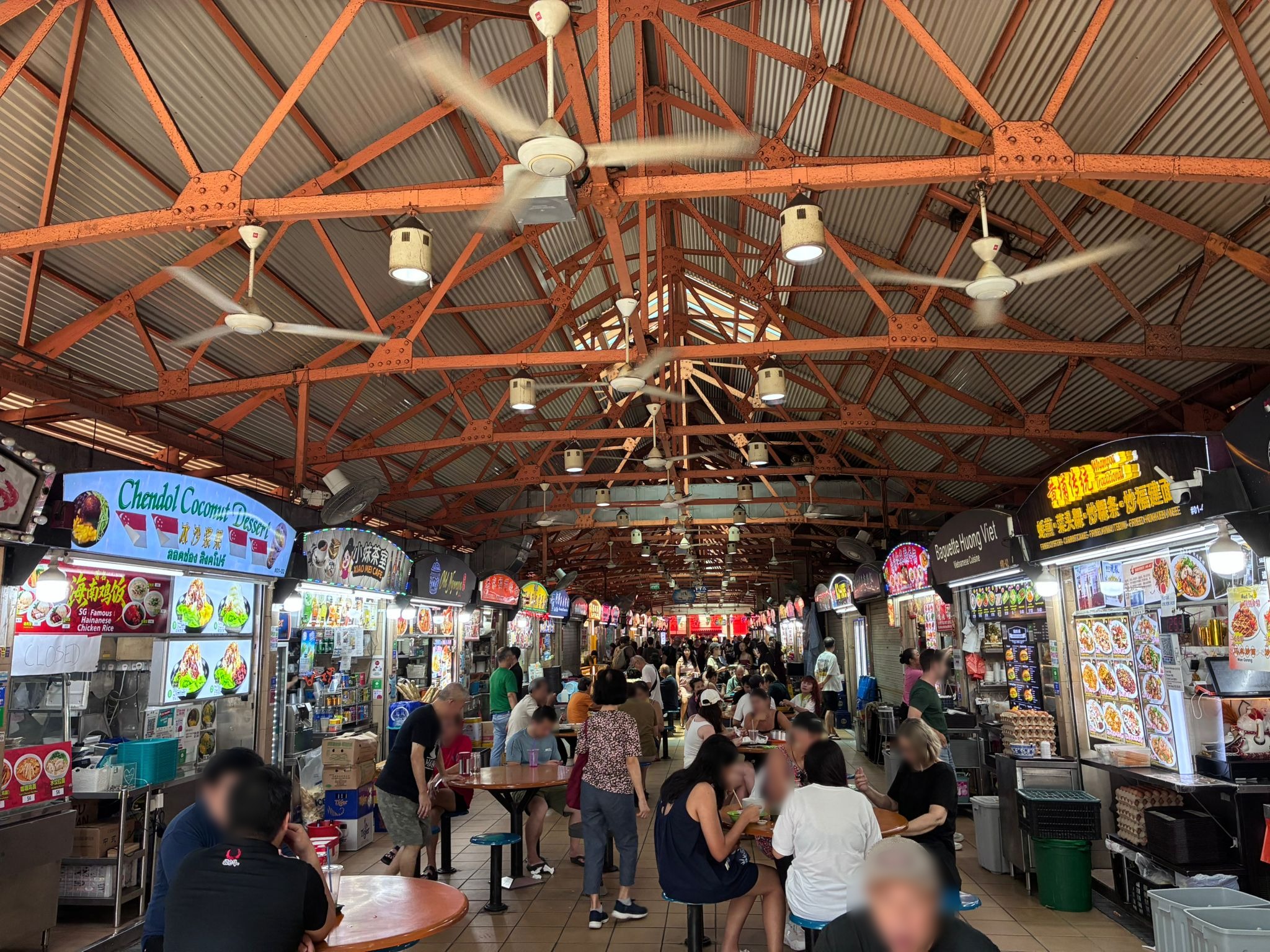 Inside Maxwell Food Centre with orange steel trusses and ceiling fans (faces blurred)