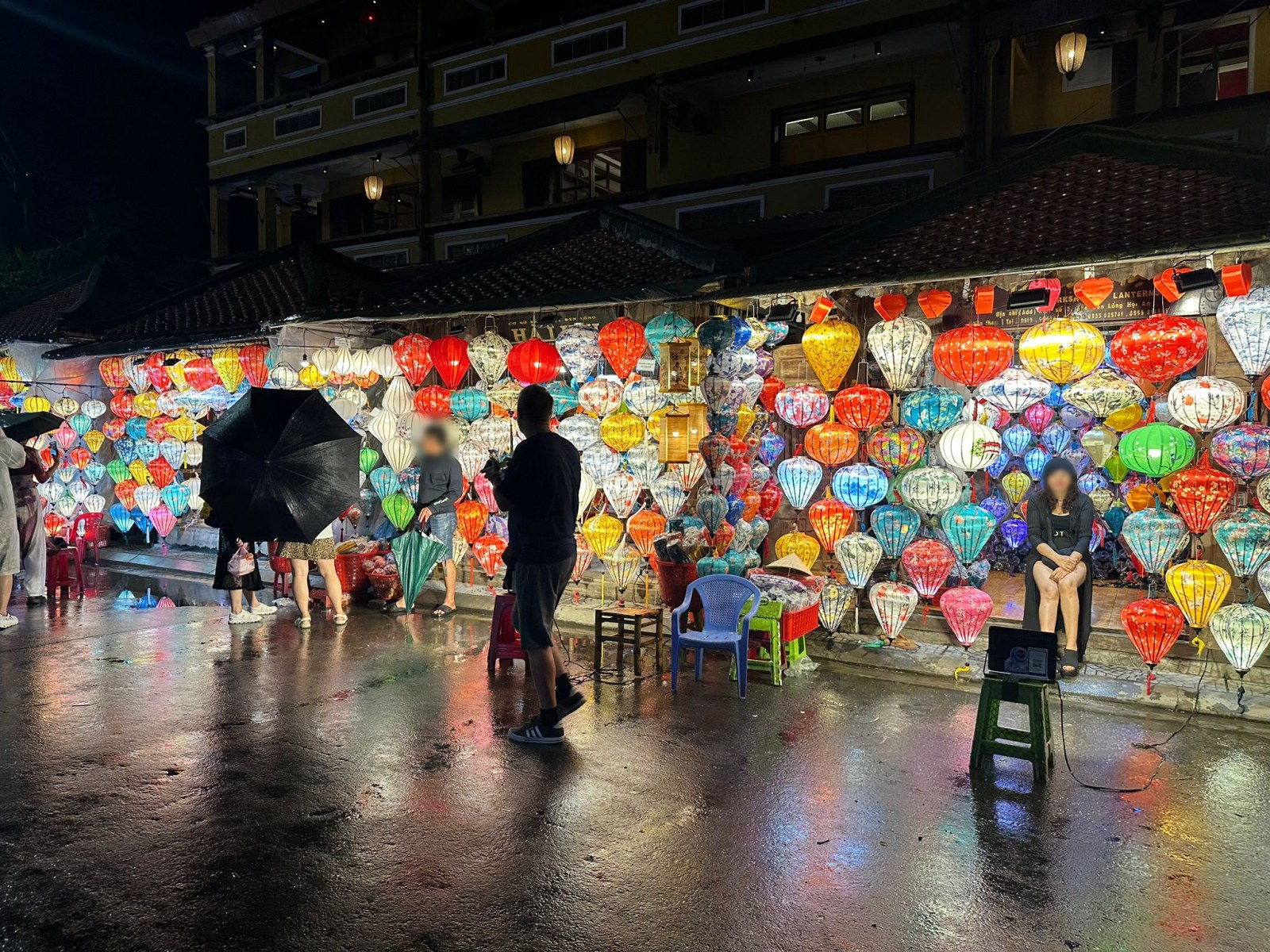 Wall of colourful silk lanterns at a shop in Hoi An old town on a rainy night, Vietnam (faces blurred)