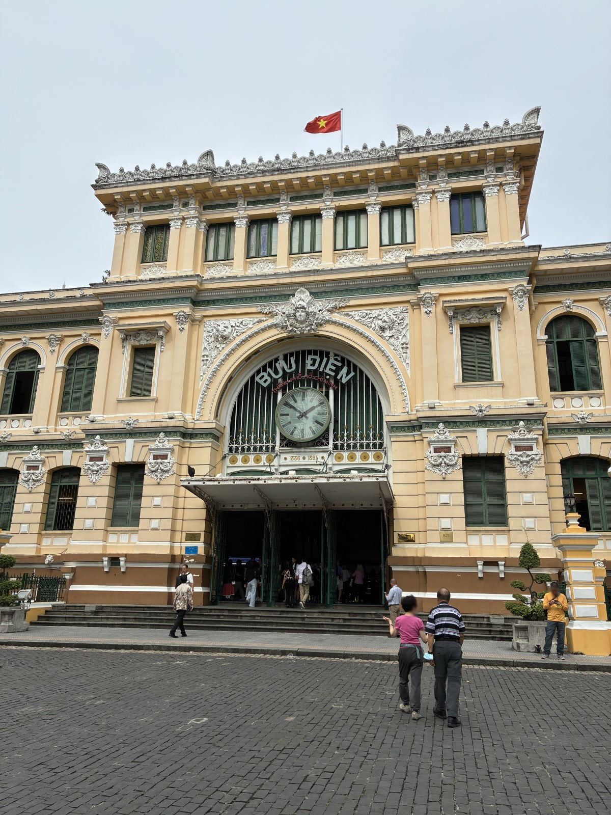 Facade of Saigon Central Post Office with clock and Vietnamese flag, Ho Chi Minh City, Vietnam (faces blurred)